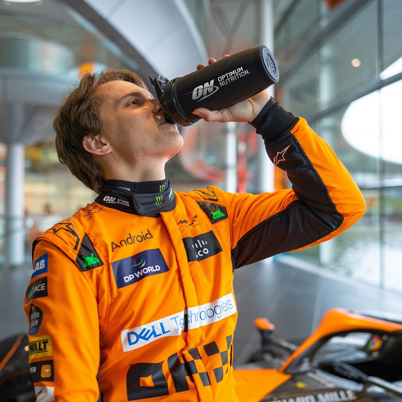 A team McLaren driver in an orange racing suit drinks from a black Optimum Nutrition shaker bottle