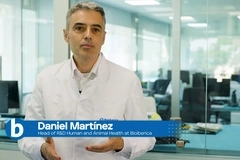 Image Man in a white lab coat stands in a glass-walled laboratory office, with a blue lower-third showing his name and role.